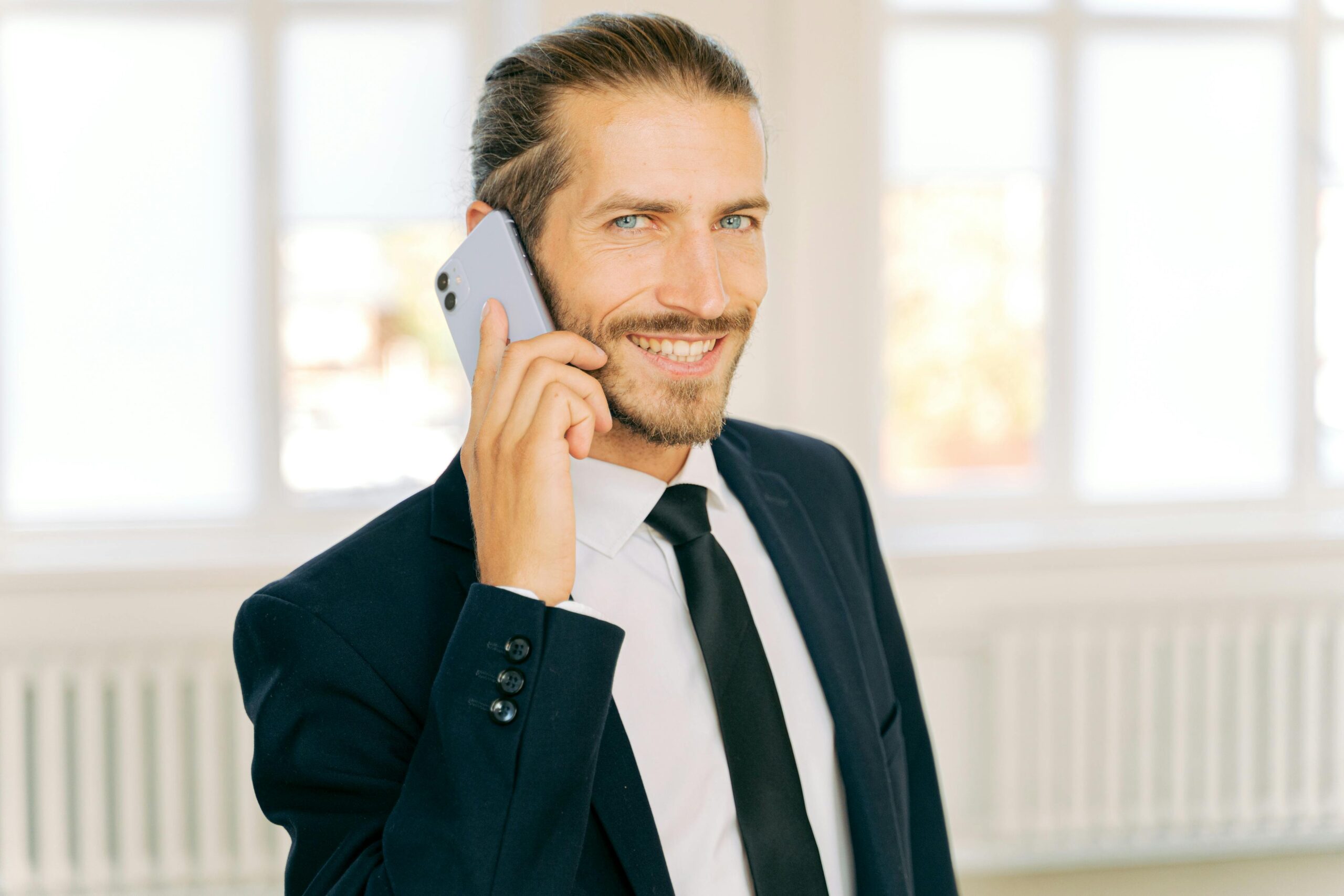 A confident businessman wearing a suit and tie, smiling while on a phone call indoors.